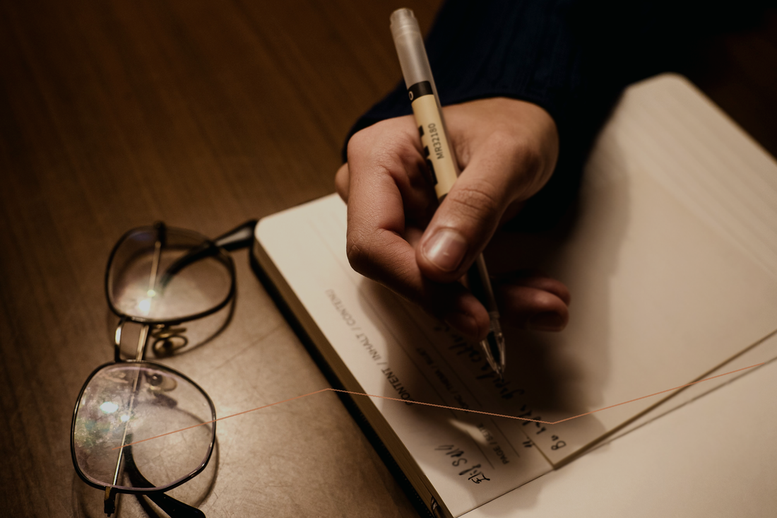 A hand writing in a notebook beside glasses in warm editorial light.