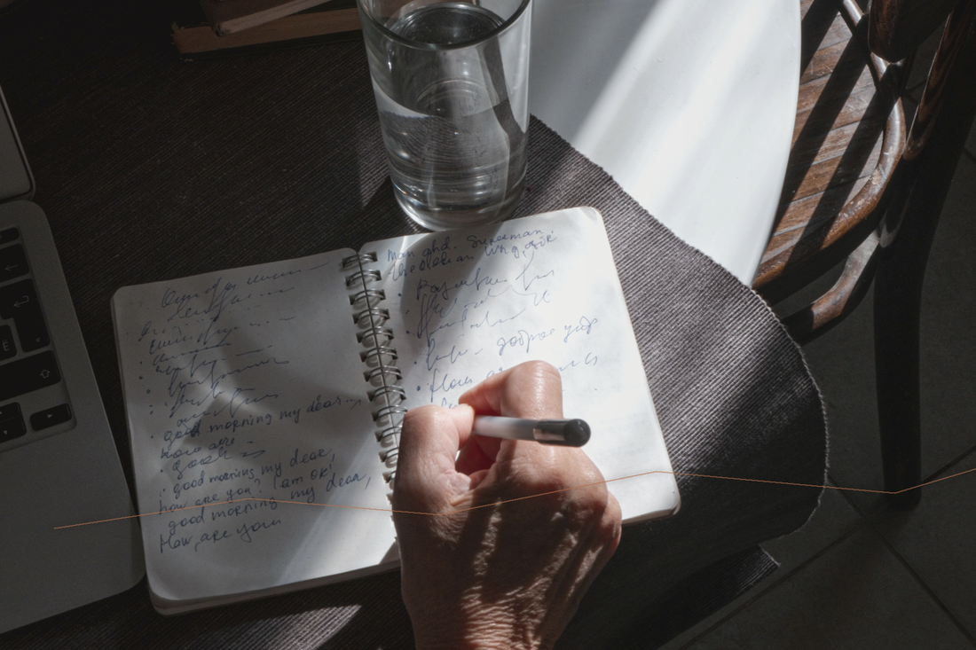 A hand writing in a notebook beside a glass of water in directional morning light.